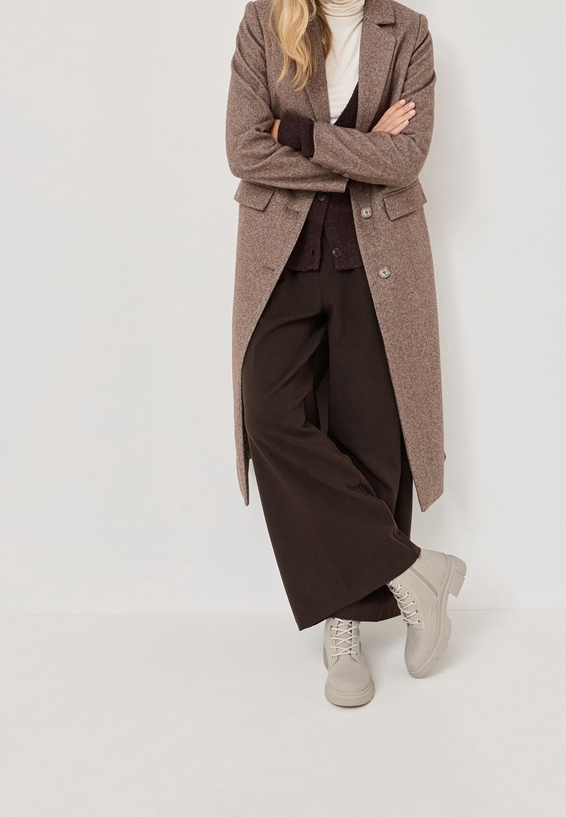 Personne debout les bras croisés, portant un long manteau marron, un pantalon large foncé, un pull à col roulé clair et des bottines beiges à lacets.