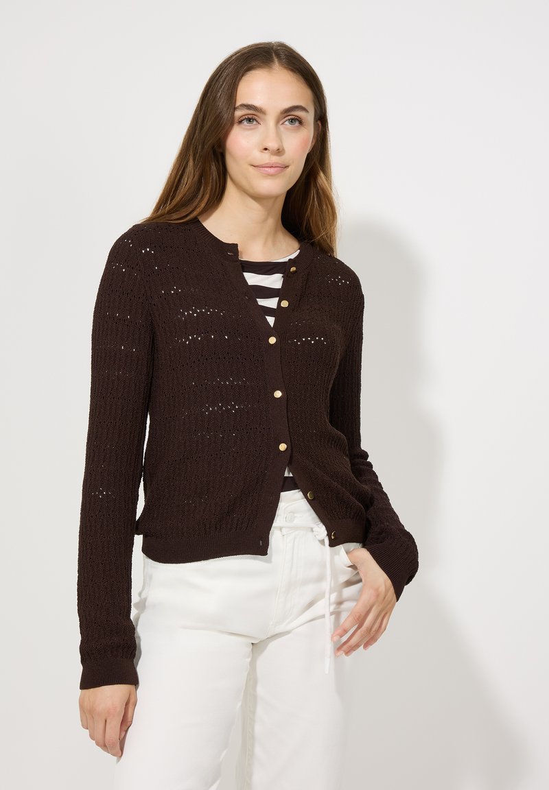 Bruine gebreide cardigan met een knoopsluiting, voorzien van een textuurpatroon en geribde manchetten, gedragen over een gestreept shirt en witte broek.