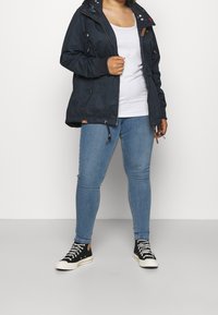 Marineblå vandtæt jakke med hætte, udstyret med snoretræk og dekorative detaljer, kombineret med lyseblå skinny jeans og sorte høj-top sneakers.