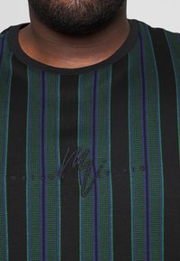 Hombre vistiendo una camisa negra con rayas verticales verdes y azules y texto negro bordado en el pecho.