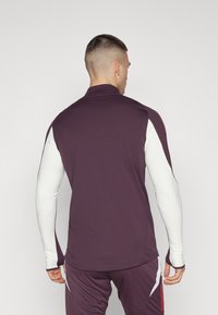 Maglietta sportiva a maniche lunghe con corpo viola scuro, maniche bianche, colletto alto e dettagli delle cuciture minimi. Tessuto morbido e elasticizzato.