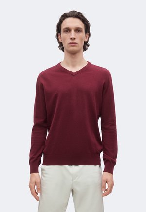 Homme portant un pull bordeaux à manches longues avec col en V et un pantalon beige clair, debout devant un fond clair uni.