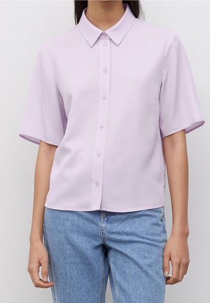 Kobieta ubrana w jasnofioletową koszulę z krótkim rękawem zapinaną na guziki, w połączeniu z jasnoniebieskimi dżinsami, na jednolitym tle.