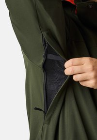 Zielona kurtka z bocznym zamkiem odsłaniającym siatkową kieszeń. Regulowany sznurek w dolnej części i gładka struktura materiału.