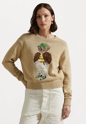Beige knitted sweater with a round neck, featuring an embroidered bear in a green cap, white pants, and sneakers. Text and details visible.