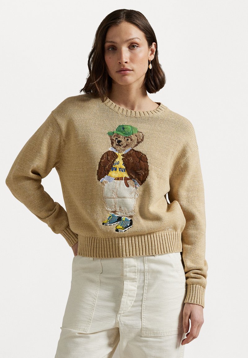 Beige gebreide trui met een ronde hals, voorzien van een geborduurde beer in een groene pet, witte broek en sneakers. Tekst en details zichtbaar.