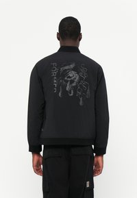 Black bomber jacket with a matte finish, featuring a large graphic print on the back, text, and a ribbed collar and cuffs. Casual fit.
