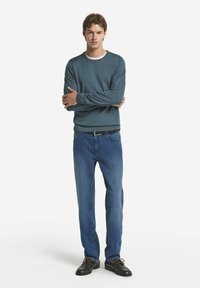 Blå långärmad tröja med vit kant, kombinerad med ljusblå jeans och svarta skor med bruna snören, stående mot en vit bakgrund.