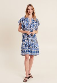 Blaumusterndes Kleid mit Rüschensäumen und V-Ausschnitt, das aufwendige Blumendesigns und einen Saum mit verzierten Details aufweist. Getragen mit schwarzen Sandalen.