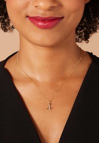 Gouden ketting met een dunne schakel en een kleine, zilveren letter "A" hanger. Huidskleur is natuurlijk; top is zwart met een V-hals ontwerp.