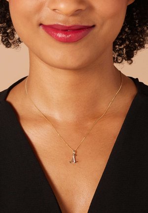 Gouden ketting met een dunne schakel en een kleine, zilveren letter "A" hanger. Huidskleur is natuurlijk; top is zwart met een V-hals ontwerp.