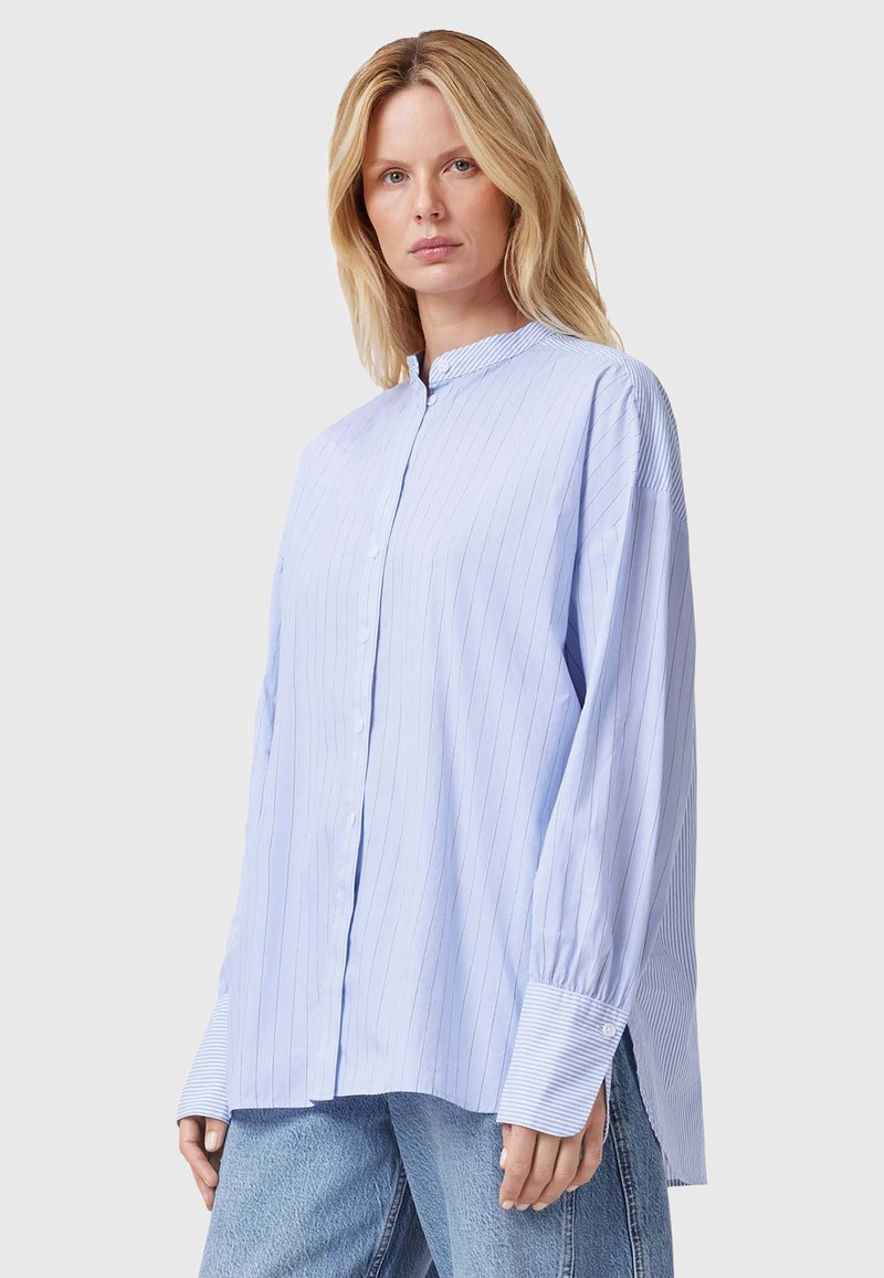 Světle modrá pruhovaná košile s button-up zapínáním, s band límcem, nadměrnými rukávy a kontrastními pruhovanými manžetami. Nosená přes džíny.