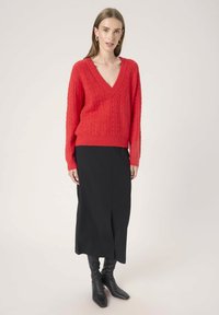 Woman wearing a red V-neck cable knit sweater, black midi skirt with front slit, and black heeled boots, standing against a plain background.