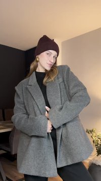 Szürke gyapjúkeverék túlméretezett kabát széles hajtókával és két gombbal, sötét sapkával párosítva. Simább textúra, laza szabás, elöl zsebek láthatók.