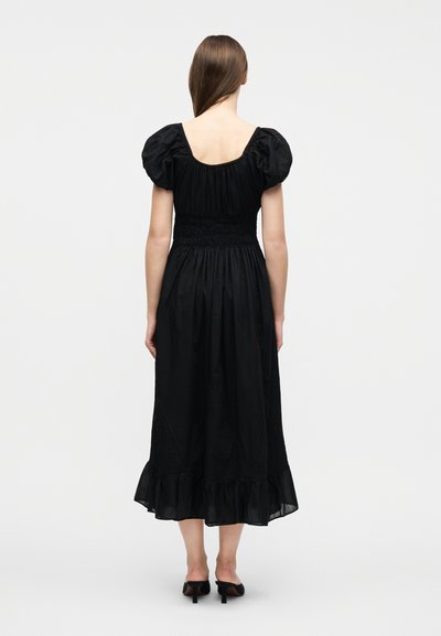 Γυναίκα με μακριά καστανά μαλλιά που φοράει μαύρο φόρεμα μίντι με φουσκωτά μανίκια και βολάν στο τελείωμα, στέκεται γυρισμένη ανάποδα, σε απλό φωτεινό φόντο.