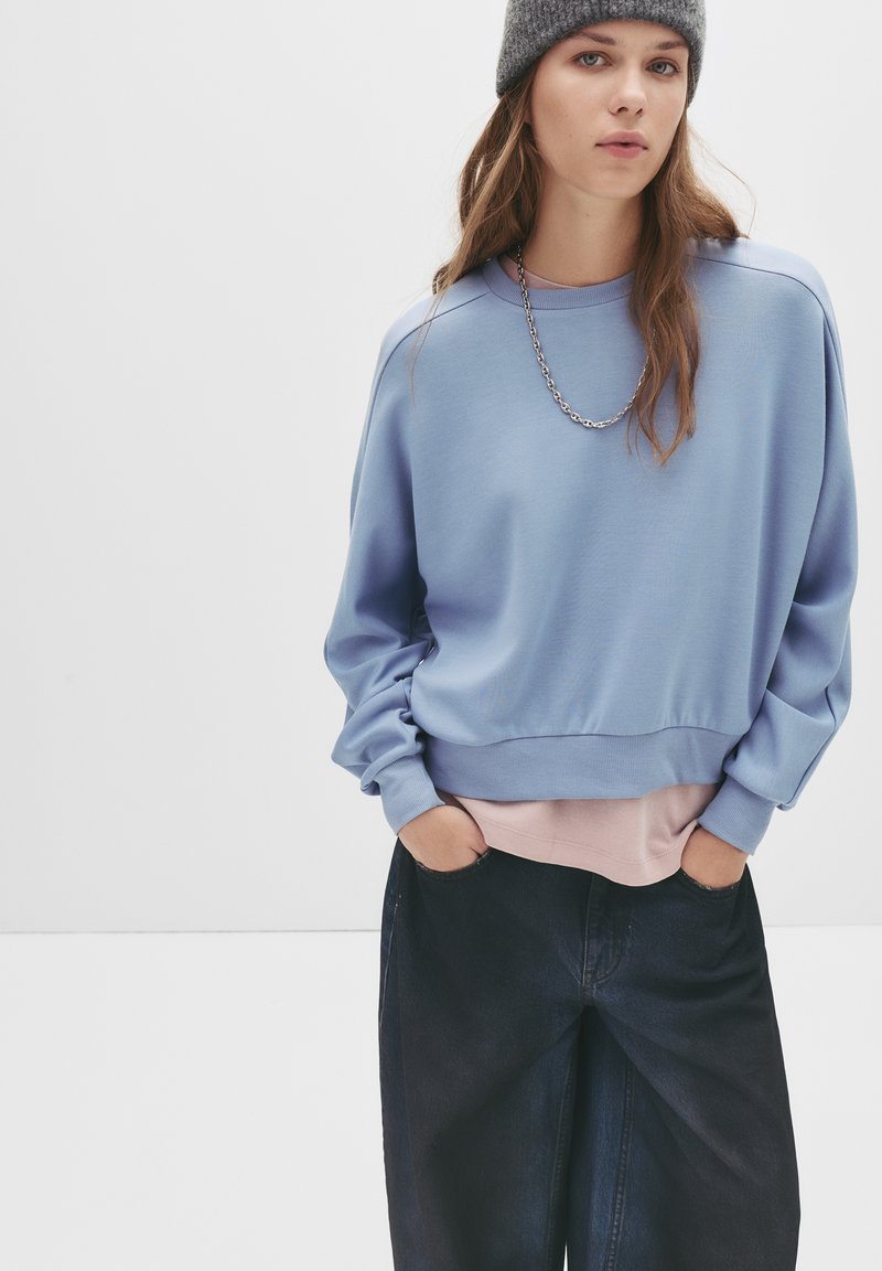 Felpa azzurra con spalle scese e orlo a costine, indossata sopra una maglietta rosa. Il modello indossa jeans scuri larghi e un berretto grigio.