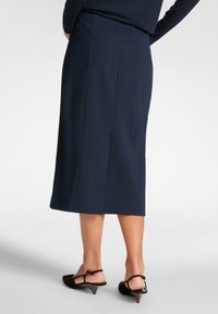 En knälång marinblå kjol i slät tyg, med en strömlinjeformad design och en diskret slits bak, parat med bruna klackskor.