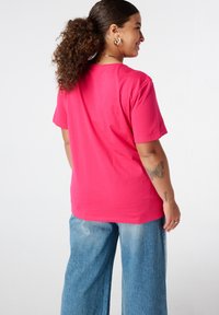 Femme aux cheveux bouclés en queue de cheval, portant un t-shirt rose et un jean bleu, de profil, souriante, montrant un tatouage de papillon sur l'avant-bras.