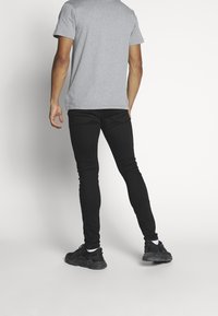 Hombre de piel marrón vistiendo una camiseta gris, jeans ajustados negros, calcetines blancos y zapatillas negras, de pie contra un fondo blanco.