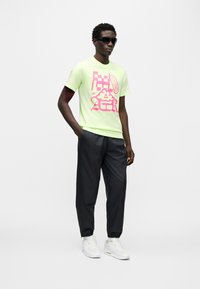 Φούξια κίτρινο μπλουζάκι με ροζ γραφικό σχέδιο, μαύρο casual παντελόνι και λευκά αθλητικά παπούτσια. Τα γυαλιά ηλίου προσθέτουν μια μοντέρνα πινελιά στην εμφάνιση.