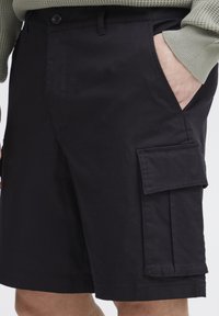 Schwarze Cargo-Shorts mit glatter Textur, seitlichen Taschen und einer Klapptasche am Oberschenkel. Hergestellt aus leichtem Material.