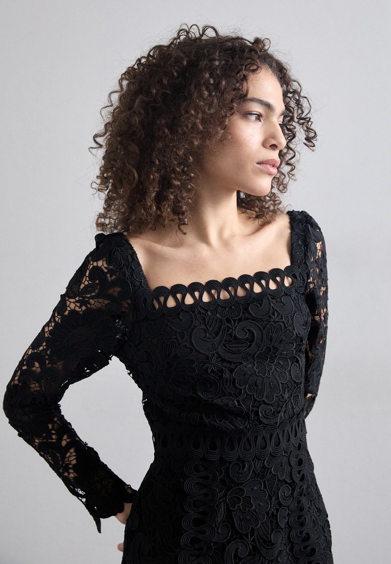 Czarna koronkowa sukienka z długimi, szczegółowymi rękawami, kwiatowymi wzorami oraz kwadratowym dekoltem z falbankami. Teksturowany, elegancki design.