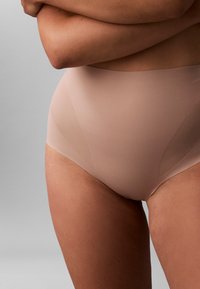 Gros plan d'une personne portant des sous-vêtements nude sans couture taille haute, bras croisés sur la poitrine, sur fond gris.