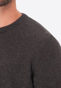 Primo piano del torso superiore di un uomo che indossa un maglione a maglia grigio scuro con scollatura a girocollo su uno sfondo chiaro e semplice.