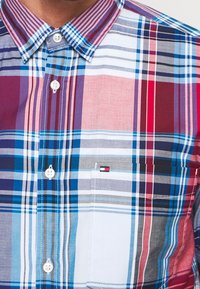 Camicia a maniche corte a quadri con motivi rossi, blu e bianchi, colletto con bottoni, tasca frontale e piccolo logo sul petto.