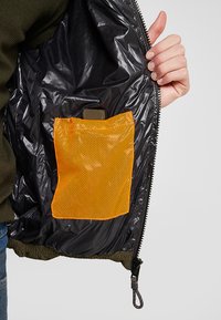 Zwarte binnenvoering van een jas met een oranje netzak. De jas heeft een rits en een gestructureerd, groen buitenmateriaal.