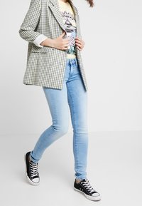 Saka v kostkovaném vzoru se zelenou a námořnickou, vrstvené přes tričko s potiskem. Slim světle modré džíny a černé tenisky dokončují outfit.