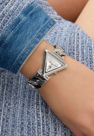 Montre triangulaire argentée avec un cadran blanc et un logo, dotée d'un bracelet en chaîne épaisse. Portée sur un poignet avec un fond en denim bleu.