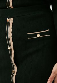 Zwart gebreid kledingstuk met een textuurpatroon, voorzien van een gouden knoopsluiting, geschulpte details en een zakaccent met beige afwerking.