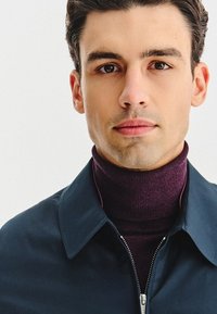 Granatowa kurtka zapinana na zamek z kołnierzem, noszona na ciemnoczerwonym golfie. Gładki materiał, dopasowany krój, minimalna widoczna metalowa biżuteria.