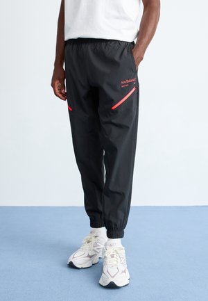 Person iført sorte New Balance træningsbukser med røde lynlåsdetailler og hvide sneakers, stående på en blå gulv mod en hvid væg.