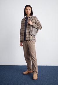 Karierter Button-Up-Hemd in Brauntönen über einem dunklen Oberteil, beigem Sweatpants mit Logo und hellbraunen Sneakers, mit einer grauen Umhängetasche.