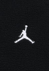 Tissu polaire noir avec un logo Jumpman blanc, représentant une silhouette en train de sauter. La texture est douce, avec une surface légèrement surélevée et duveteuse.
