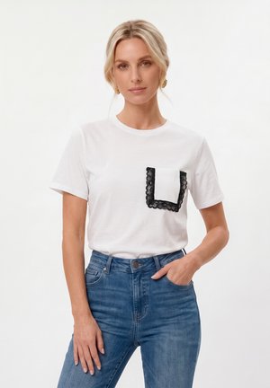 Femeie cu păr blond care poartă un tricou alb cu un buzunar cu dantelă neagră și blugi albaștri, stând cu o mână în buzunar.
