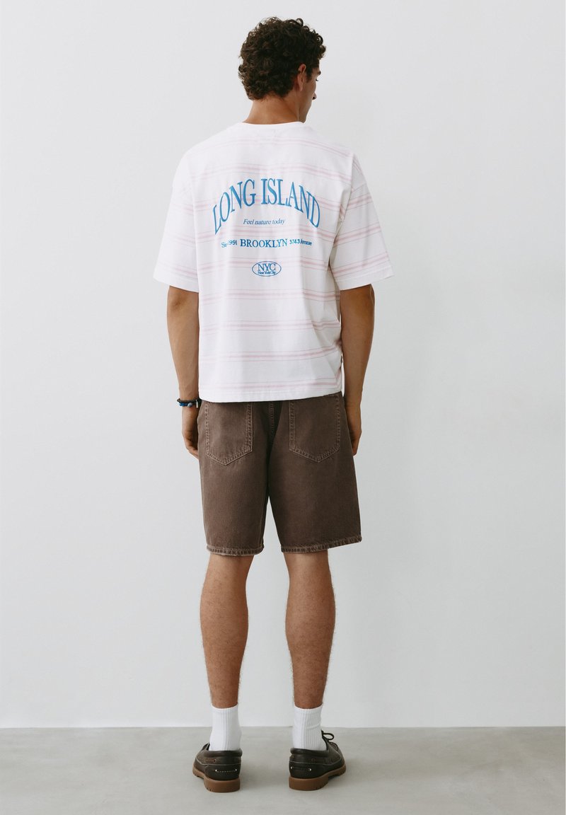 Homme debout face à un mur, portant un t-shirt blanc avec un motif Long Island, un short marron, des chaussettes blanches et des chaussures décontractées foncées.