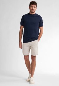 Námořnicky modrá košile s krátkým rukávem ve spojení s lehce béžovými pruhovanými šortkami. Osoba má na sobě krémové tenisky a stojí před jednoduchým světlým pozadím.