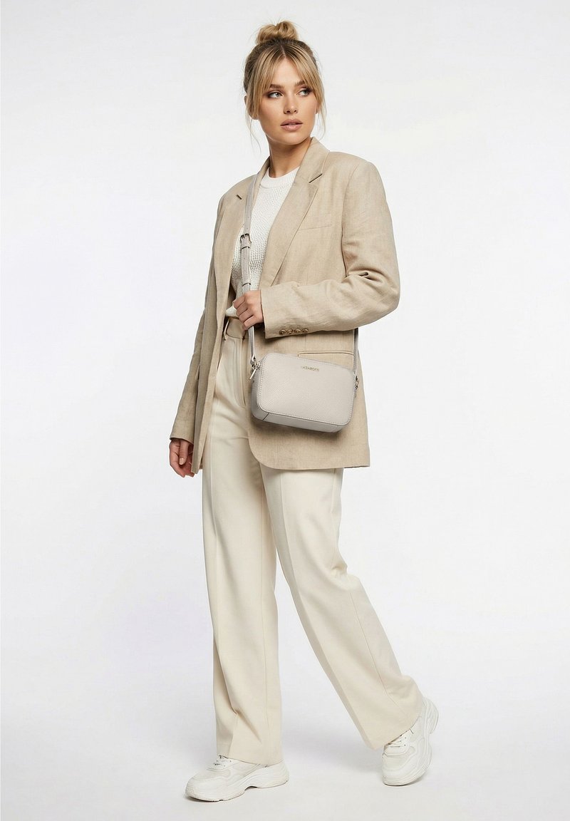 Femme aux cheveux blonds attachés en chignon, vêtue d'un blazer beige, d'un top blanc, d'un pantalon crème, de baskets blanches et d'un sac bandoulière gris clair, debout sur fond blanc.