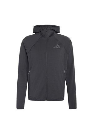 Jeune homme aux cheveux bouclés portant un sweat à capuche noir zippé et un pantalon beige, debout les mains dans les poches devant un fond uni.