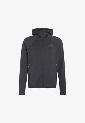 Giovane uomo con capelli ricci che indossa una felpa nera con zip e pantaloni beige, in piedi con le mani in tasca davanti a uno sfondo semplice.