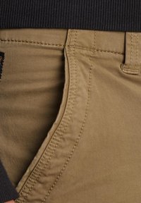 Beige katoenen broeken met een gladde textuur, voorzien van een zijzak en een contrasterende donkere ribgebreide tailleband. Zichtbare stikseldetails.