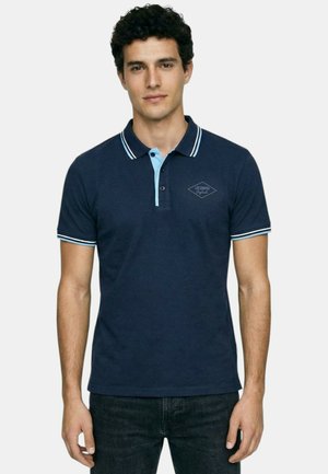 Homme portant un polo bleu marine avec une patte de boutonnage bleu clair et un col rayé, debout devant un fond uni.