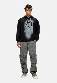 Felpa nera con un design grafico grigio, dotata di tasca frontale e cordini. Abbinata a pantaloni cargo grigi e sneakers verdi.