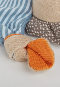 Pieds de canard en tissu doux avec toile orange attachée à des jambes de jouet portant un pantalon rayé bleu et blanc et à motifs marron, posés sur une surface blanche.