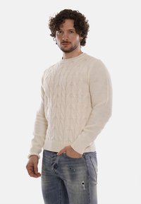 Uomo con capelli ricci che indossa un maglione a trecce color crema e jeans blu consumati, in piedi con una mano in tasca.