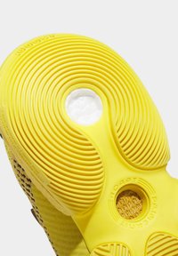 Semelle de chaussure jaune texturée avec des rainures circulaires, un coussinet blanc visible à travers un trou central, et un tissu tissé visible à travers un petit trou.