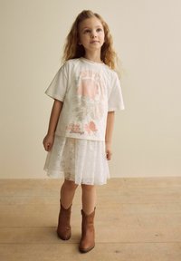 T-shirt graphique blanc avec un design floral rose et du texte, superposé avec une jupe en dentelle blanche et des bottes de cowboy brunes. Texturé et ludique.